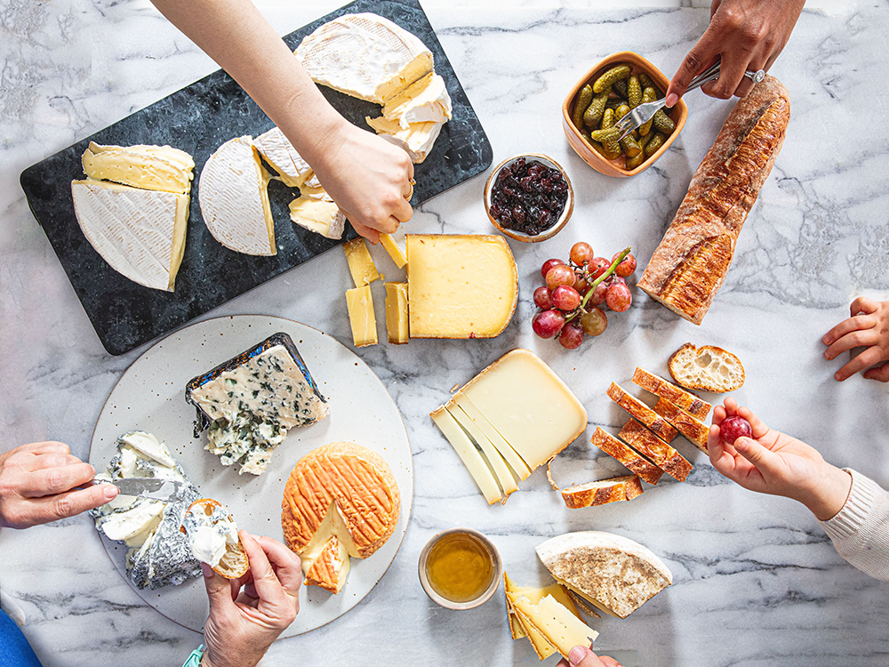 GVL A variety of cheeses, including blue, brie, and gouda, are on plates with baguette slices, grapes, pickles, and a bowl of jam. A cup of wine sits nearby as several hands reach in to share the food on a marble surface.
