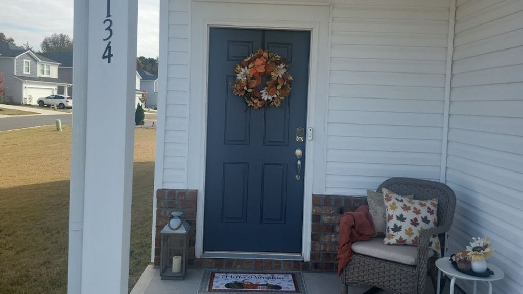 GVL A cozy porch with a blue door adorned with an autumn wreath. A wicker chair with a leaf-patterned pillow and a blanket sits nearby. A lantern is on the ground, and a small table holds a decorative pumpkin. The house number 134 is visible.