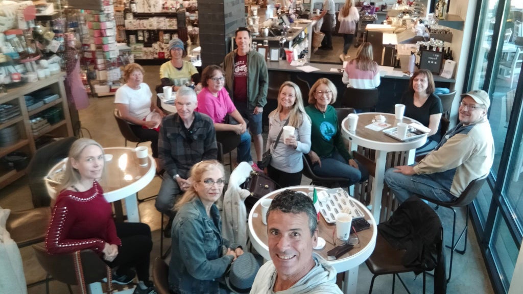 GVL A group of people are gathered in a coffee shop, sitting around tables and holding drinks. The atmosphere is lively, with shelves of products visible in the background. A man in the foreground takes a selfie with the group.