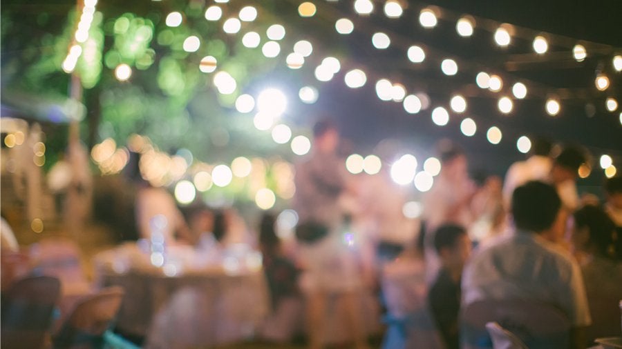 GVL A blurred image of an outdoor event with string lights glowing above. People are seated at tables, creating a warm, festive atmosphere. The setting appears to be a celebration, such as a wedding or party, held in the evening.