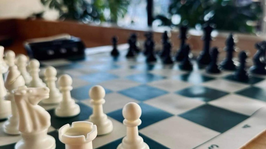GVL A chessboard is set up for a game, displaying white and black pieces in starting positions. The focus is on the white pieces in the foreground, with blurred greenery in the background.