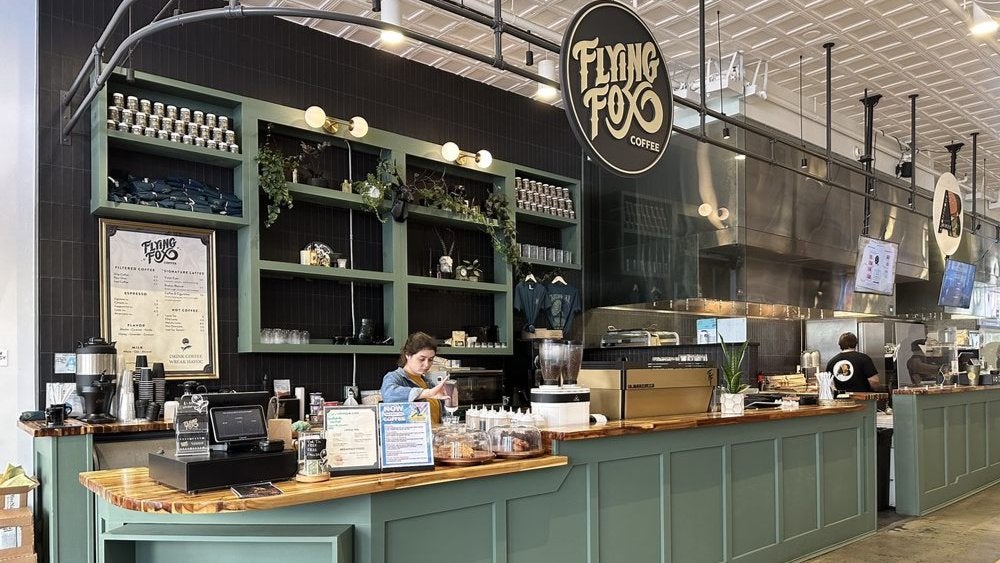 GVL A woman works behind the counter at Flying Fox Coffee, a café with a green-themed interior. Shelves with plants and coffee bags are displayed above the counter. The café menu and equipment like espresso machines are visible.