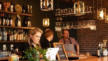 GVL A cozy bar scene with three people. A woman is using a tablet at the counter, while two men in aprons are behind the bar. Shelves with bottles and hanging glasses are visible, with warm lighting creating an inviting atmosphere.