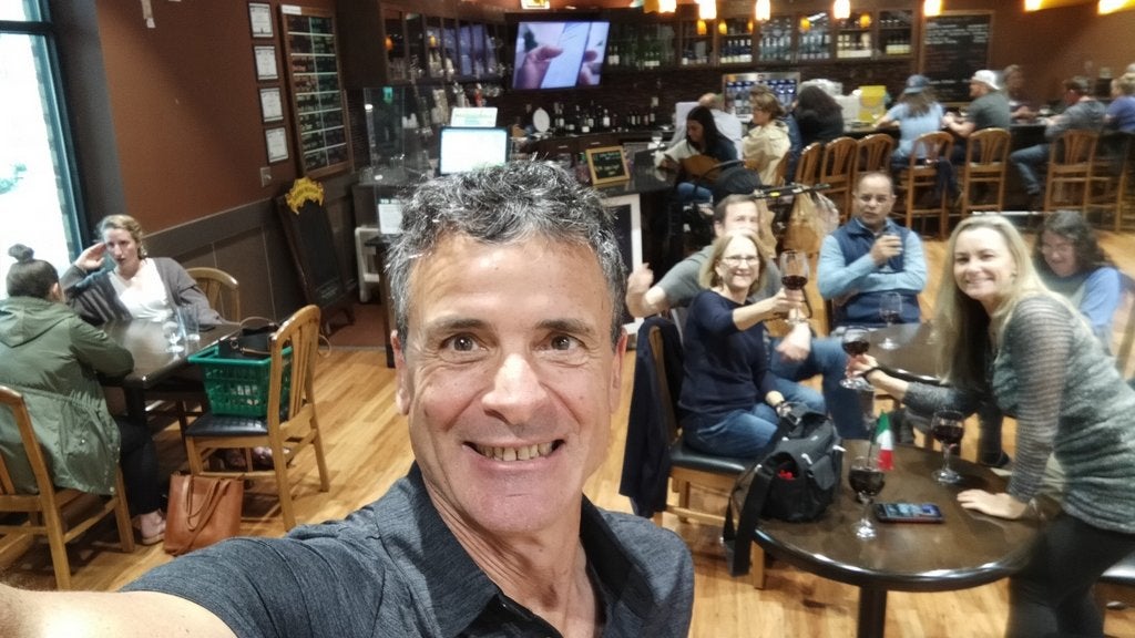 GVL A smiling man takes a selfie in a lively café or restaurant. People sit at wooden tables in the background, chatting and holding drinks. The atmosphere is warm and welcoming. A television is on the wall, and wine bottles are displayed behind a counter.