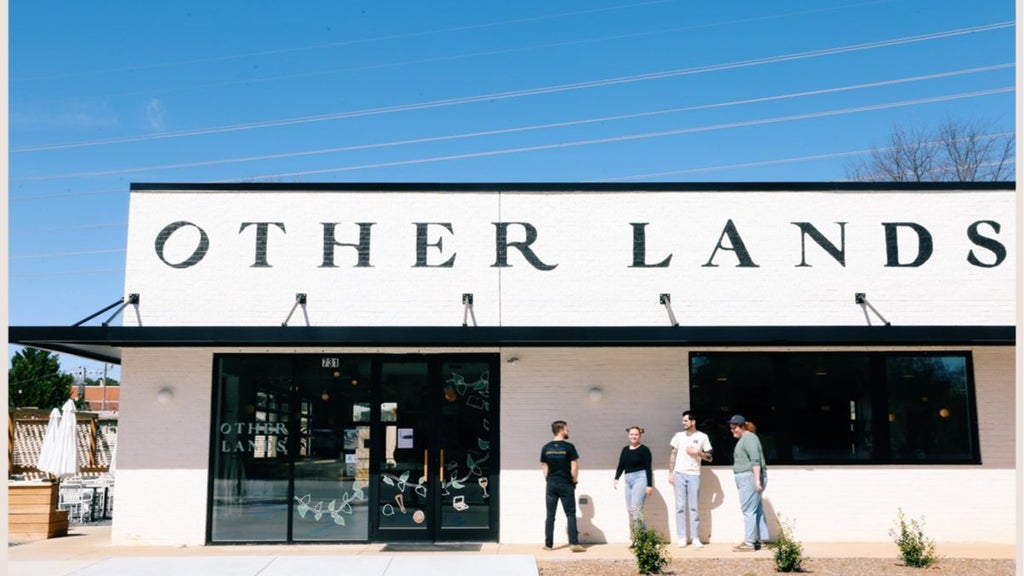 GVL Street view of a building with a white facade and OTHER LANDS in large black letters. Four people stand in conversation near the entrance. The building has a modern design with large windows and a small planting area in front.