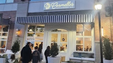 GVL A group of people stands outside Carmellas Café & Dessert Bar in the evening. The café has a striped awning and is warmly lit. Small potted trees decorate the entrance area.
