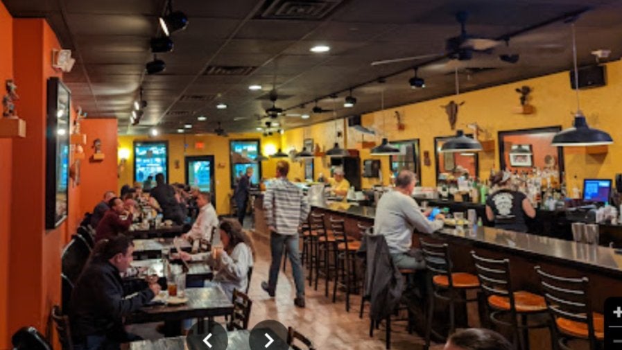 GVL A cozy restaurant interior featuring a bar on the right with patrons seated on barstools, tables filled with diners on the left, warm lighting, and walls adorned with decorative items. The atmosphere is lively with people enjoying food and drinks.