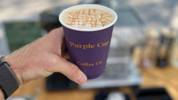 GVL A hand holds a purple coffee cup with Purple Cup Coffee Co written on it. The cup contains a yellowish-brown beverage topped with a caramel drizzle pattern. The background is blurred, displaying outdoor elements.