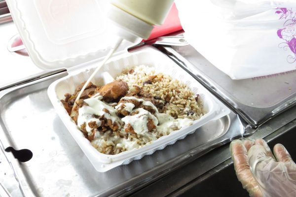 GVL A person wearing gloves is drizzling white sauce over a takeout container brimming with grilled chicken and rice. The container sits on a metal counter in a bustling food cart, reminiscent of the lively atmosphere found near Yankee Stadium.