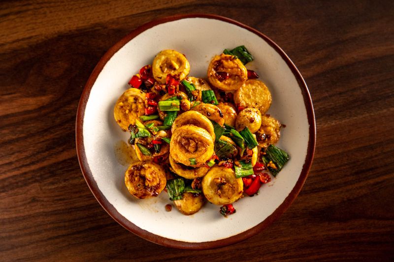 GVL A white plate with a brown rim contains a dish of round, pan-seared tofu pieces topped with a savory sauce. Reminiscent of NYCs vibrant Chinese restaurants, its accompanied by chopped green onions and red bell pepper pieces, resting on a wooden tabletop.