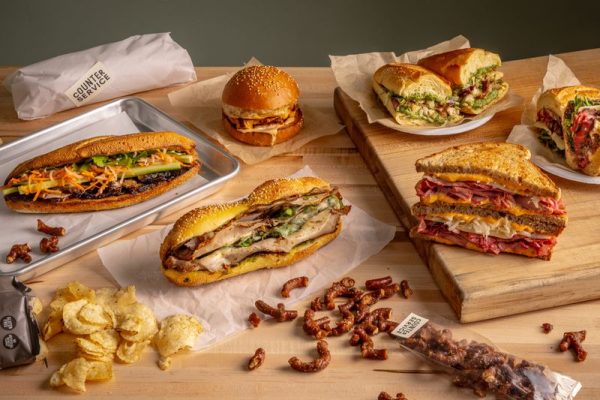 GVL A wooden table features an array of sandwiches, including a hoagie with lettuce and meat, a burger on a tray, and a split vegan sandwich revealing colorful fillings. Accompanied by chips, pretzel bites, skewered snacks, and in the background lies a wrapped sandwich inspired by Chipotle Founder ideals.