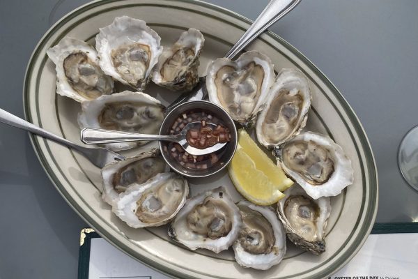 GVL A plate of the best oysters on the half shell is perfectly arranged in a circle, with a zesty bowl of sauce in the center and a lemon wedge on the side. Two metal spoons rest nearby, inviting you to indulge during New York Citys happiest hours. The menu border peeks from below.