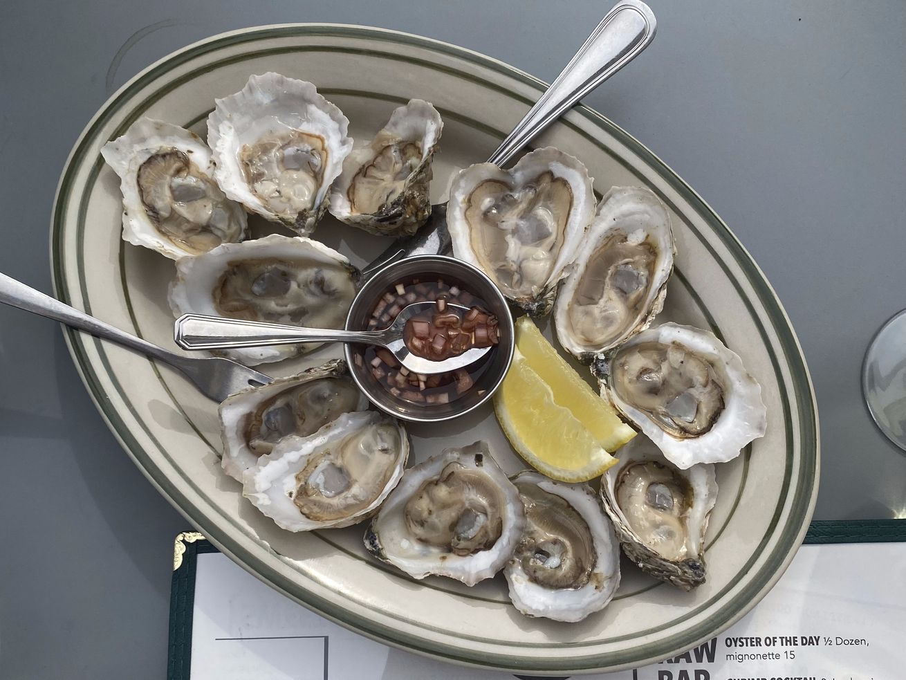 GVL A plate of the best oysters on the half shell is perfectly arranged in a circle, with a zesty bowl of sauce in the center and a lemon wedge on the side. Two metal spoons rest nearby, inviting you to indulge during New York Citys happiest hours. The menu border peeks from below.