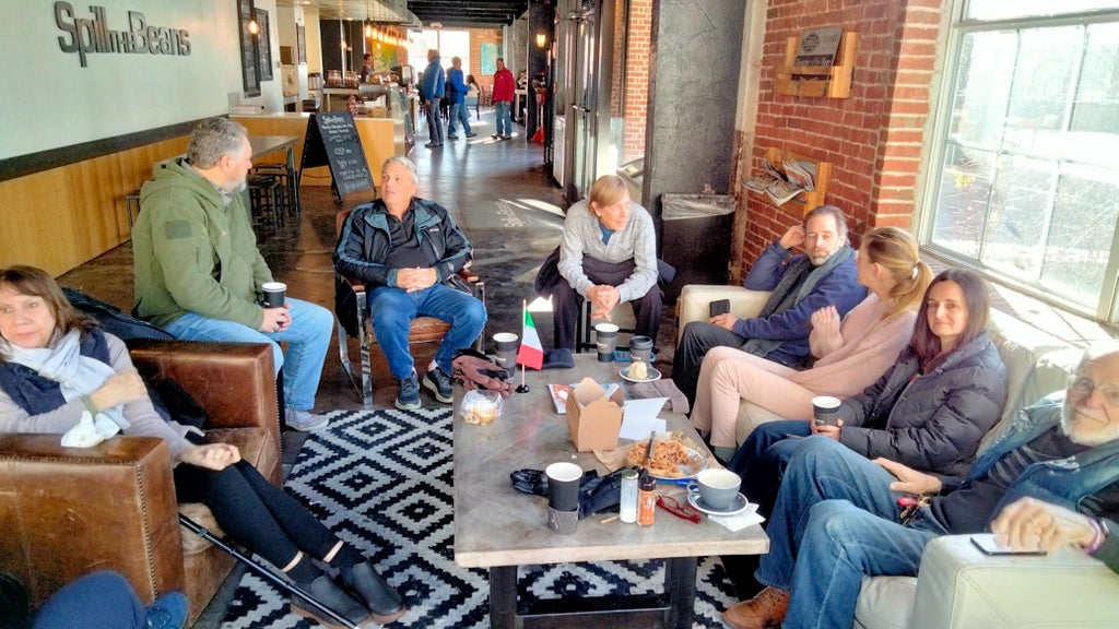 GVL A group of people sit and chat in a cozy coffee shop with brick walls and a patterned rug. Some hold cups, and a table in front of them has food containers and drinks. Sunlight streams in through large windows in the background.
