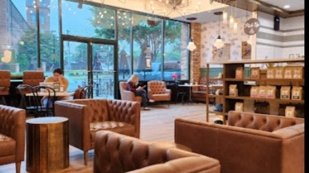 GVL A cozy cafe with brown leather chairs and a wooden floor. Large windows let in natural light. A person is sitting and reading near the window, while another sips coffee at a table. Shelves display coffee bags and decor. Warm lighting creates a relaxed ambiance.