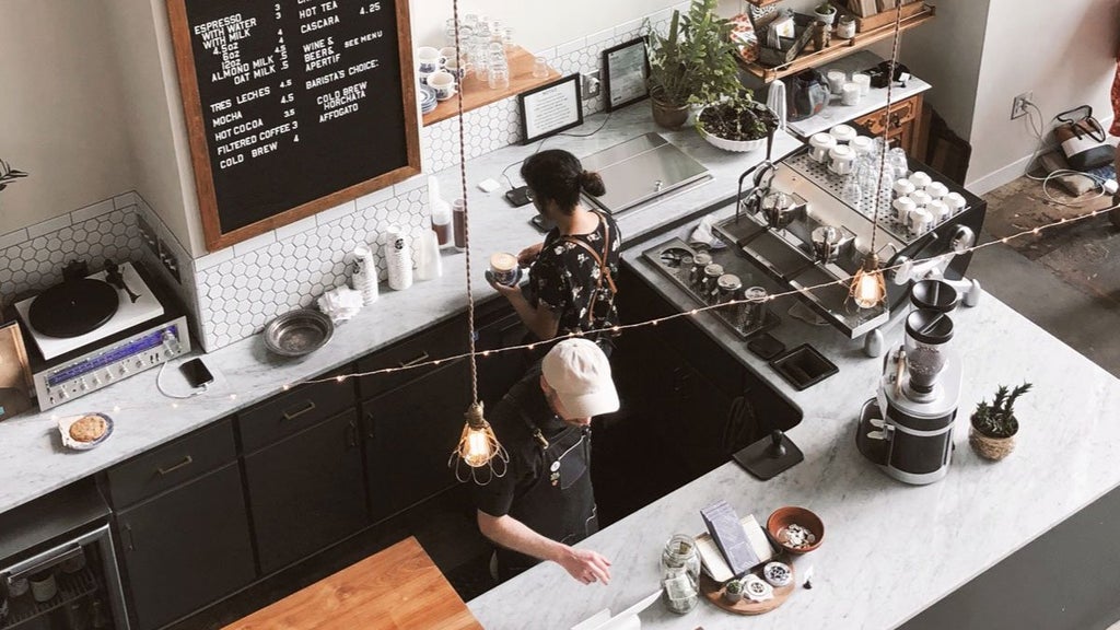 GVL Aerial view of a cozy coffee shop with two baristas preparing drinks. The counter has a coffee machine, utensils, and plants. A menu is visible on the wall, and soft lighting adds a warm ambiance.