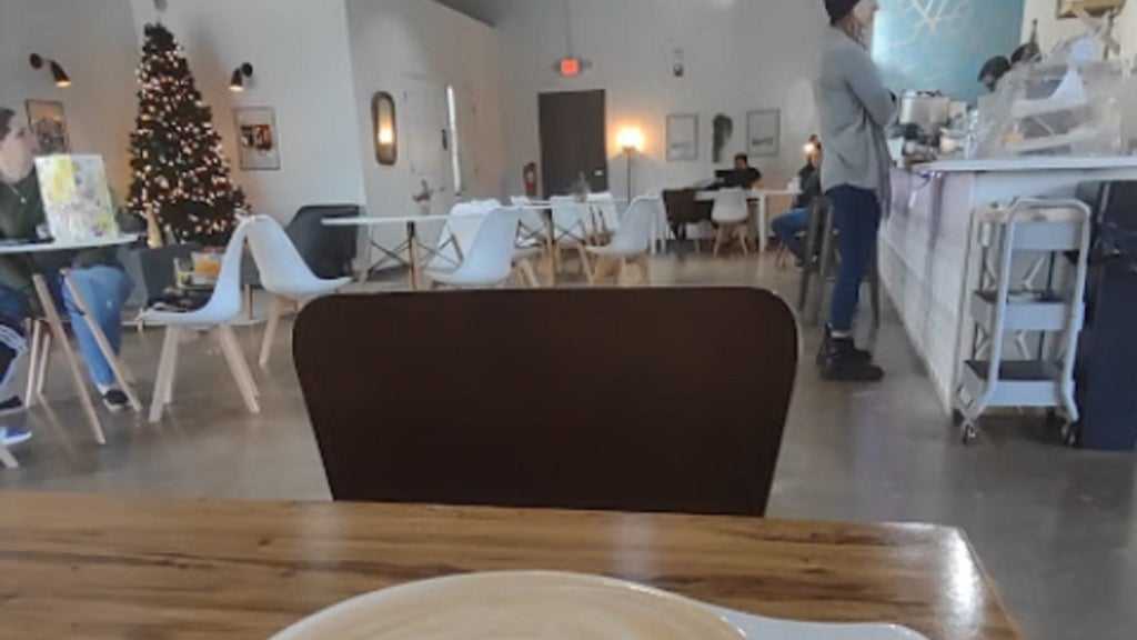 GVL A cozy café with a Christmas tree in the corner. People are seated at tables, and a person is standing near the counter. The foreground shows a wooden table with a latte on it.