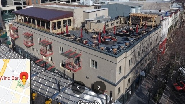 GVL Aerial view of a multi-story building with a rooftop terrace featuring seating and umbrellas. Adjacent streets and buildings are visible. A small inset in the bottom left shows a map location pinpoint labeled Wine Bar.