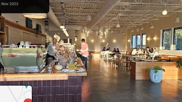 GVL A spacious, sunlit café with high ceilings and large windows. People are sitting at tables and a barista is behind the counter with a coffee machine and autumn-themed decorations.