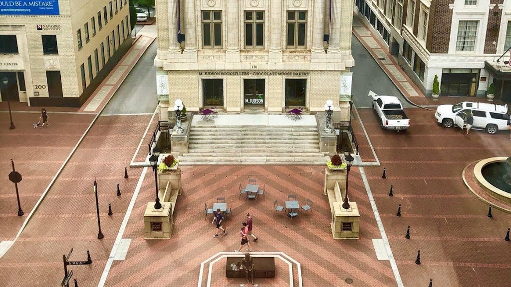 GVL Aerial view of a public square with brick pavement and geometric patterns. A few people walk across the space, surrounded by historic buildings. Stairs lead up to a grand entrance with columns flanked by stone planters. Cars are parked nearby.