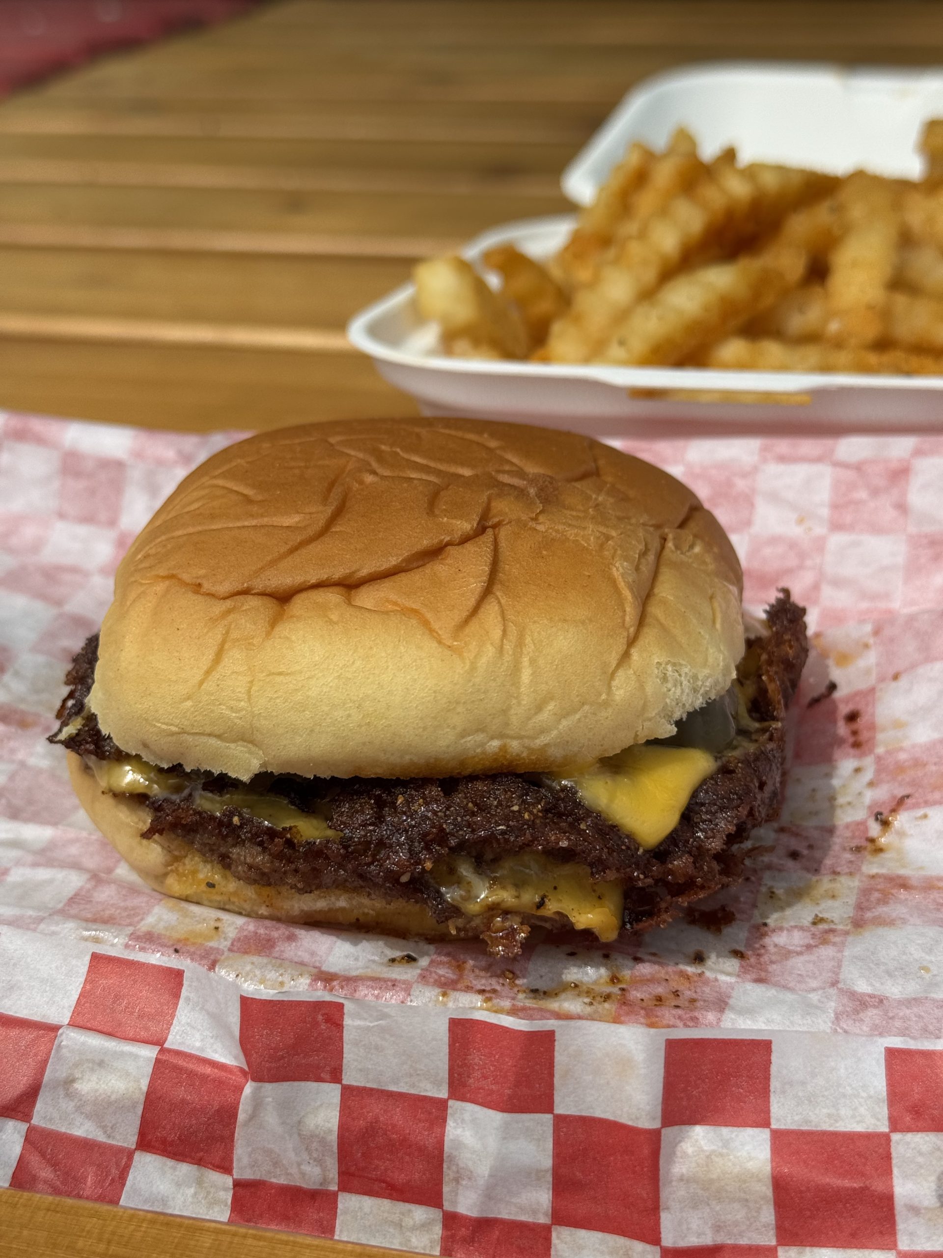 GVL A cheeseburger with a toasted bun and melted cheese sits on red-and-white checkered paper, with a container of crinkle-cut fries in the background on a wooden table.