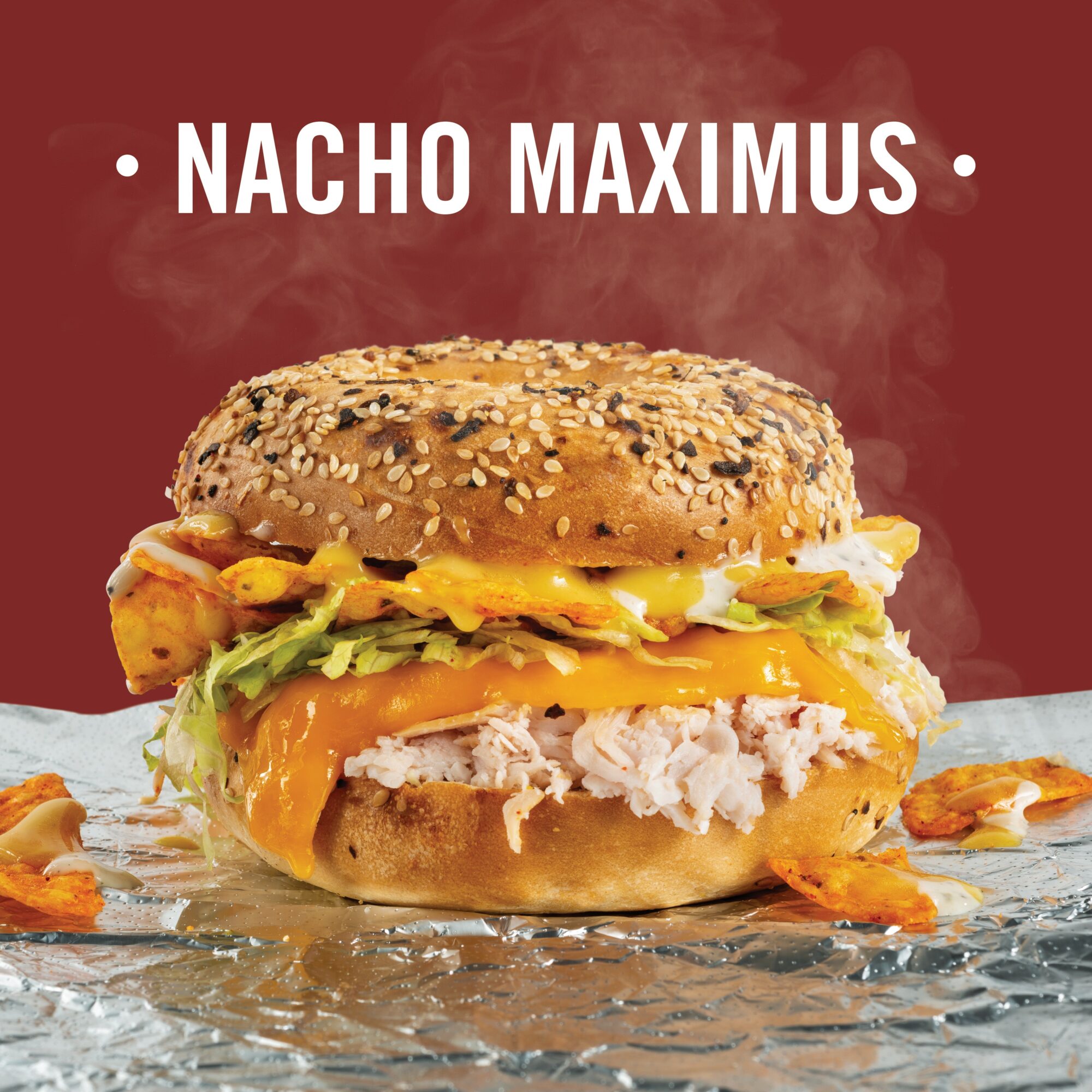 GVL A sesame seed bagel sandwich filled with shredded meat, melted cheddar cheese, lettuce, nacho chips, and sauce sits on foil, with the words “NACHO MAXIMUS” above it on a red background.
