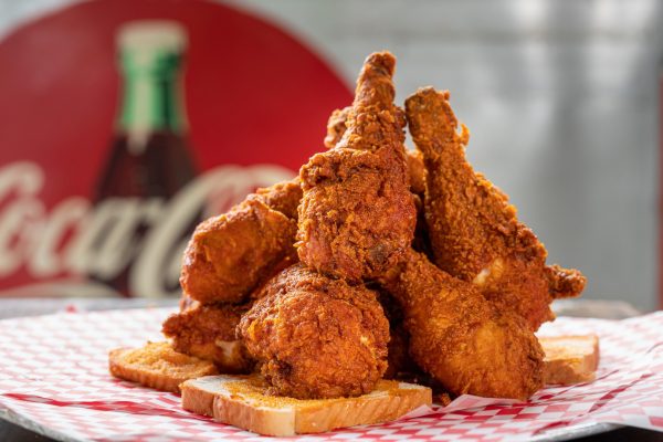GVL A pile of crispy fried chicken drumsticks stacked on slices of white bread, served on red and white checkered paper with a blurred Coca-Cola sign in the background.