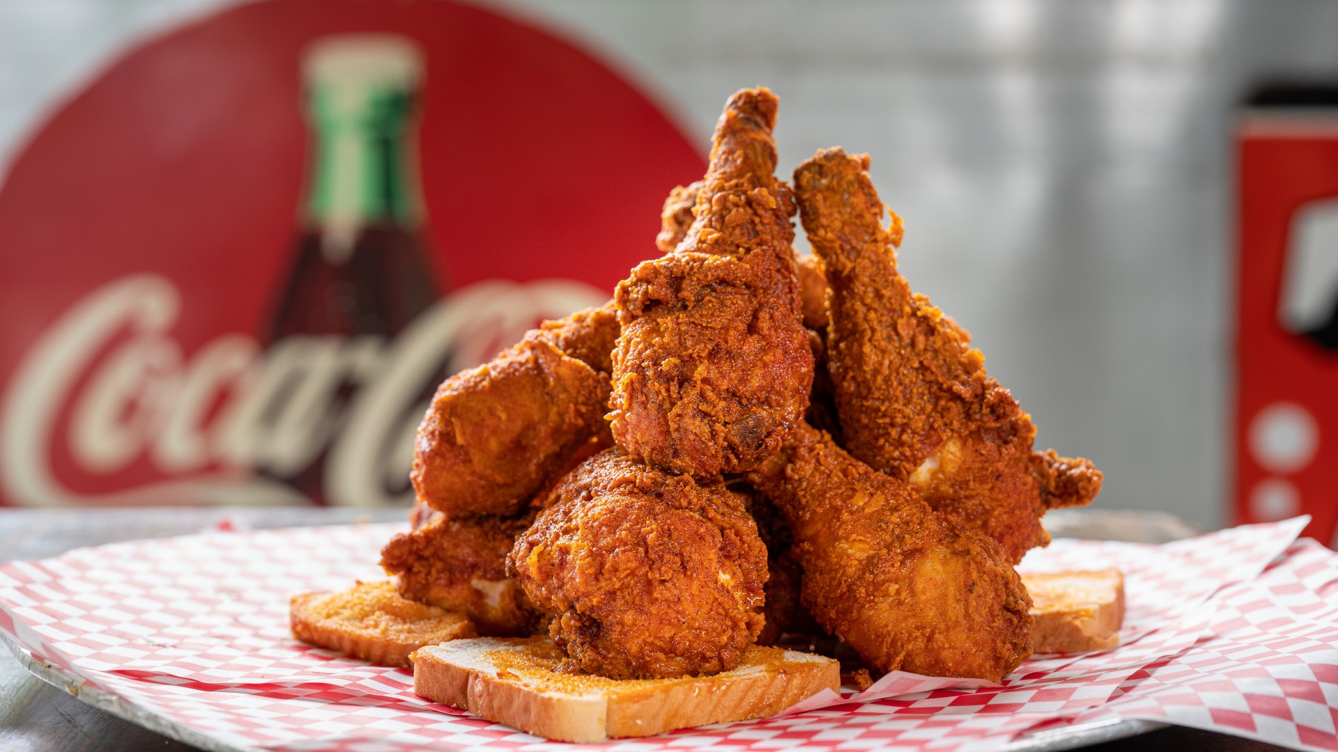 GVL A pile of crispy fried chicken drumsticks stacked on slices of white bread, served on red and white checkered paper with a blurred Coca-Cola sign in the background.