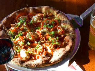 GVL A pizza topped with green onions, red peppers, and sauce sits on a plate next to a glass of City Juice and beer, all on a wooden table in sunlight—a scene that could’ve come from John Malik’s kitchen.
