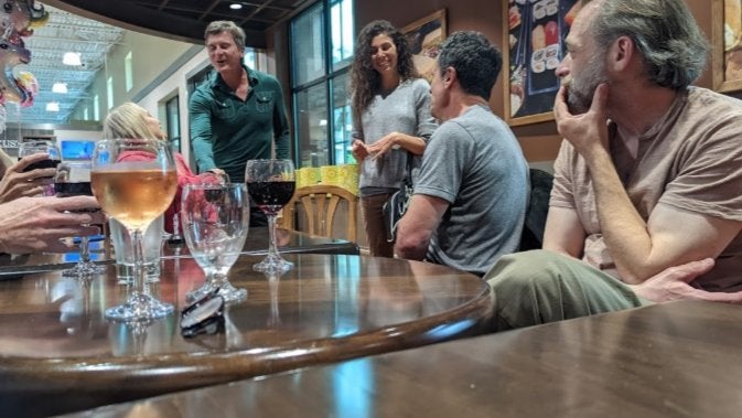 GVL A group of people sit and stand around a wooden table in a restaurant or café, talking and enjoying drinks. Several glasses of wine and water are on the table. The atmosphere appears casual and friendly.