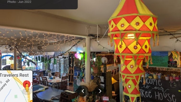 GVL A colorful, triangular yellow and red hanging lantern is in the foreground of a cozy, decorated café with string lights, eclectic furniture, and a chalkboard sign that reads, Welcome to our Tree House.