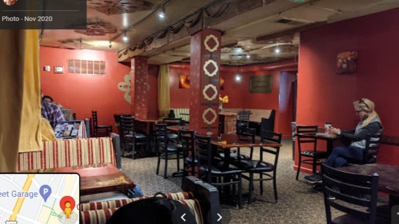 GVL Cozy café with red walls, patterned ceiling, and scattered tables and chairs. A few people are seated; one woman sits alone at a table using a phone. Warm lighting creates a relaxed atmosphere. A map is partially visible in the bottom left corner.