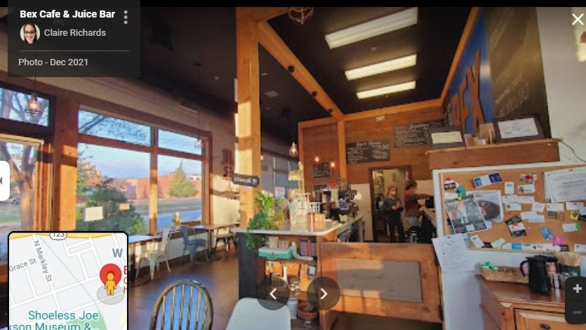 GVL A cozy cafe interior with wooden decor, large windows showing sunlight outside, a counter with menu boards, and a bulletin board on the right wall. A few people are behind the counter. A small map is visible in the corner.