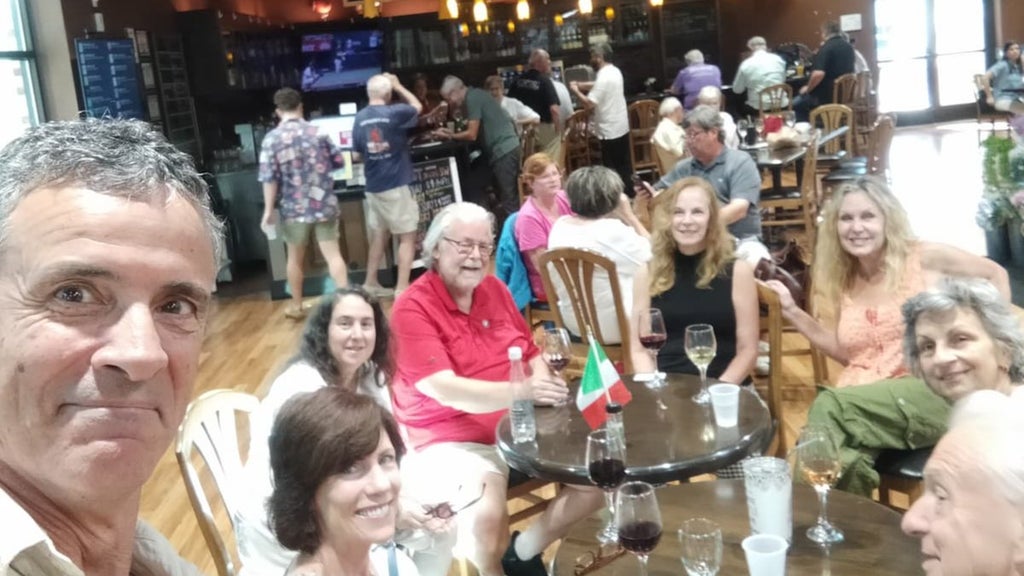 GVL A group of people sit around a table in a lively, well-lit restaurant or bar, smiling at the camera. Drinks and a small Italian flag sit on the table; other patrons can be seen in the background.