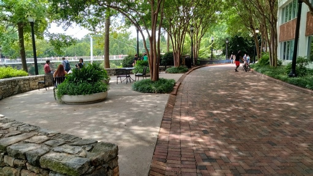 GVL A tree-lined walkway with stone and brick paths, benches, and people sitting or walking on a sunny day. Buildings and a river are visible in the background.
