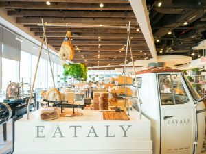 GVL A bright, modern market interior at Eataly NYC features a white counter labeled EATALY, displaying cured meats, a deli slicer, and wooden cutting boards—a glimpse of expansion with a small white vehicle beside the counter.