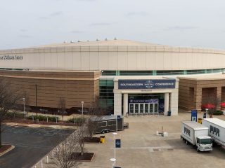 GVL A large, round arena building with a sign reading Southeastern Conference above the entrance. Several trucks are parked outside Bon Secours Wellness Arena, and a few leafless trees surround the area on a cloudy day.