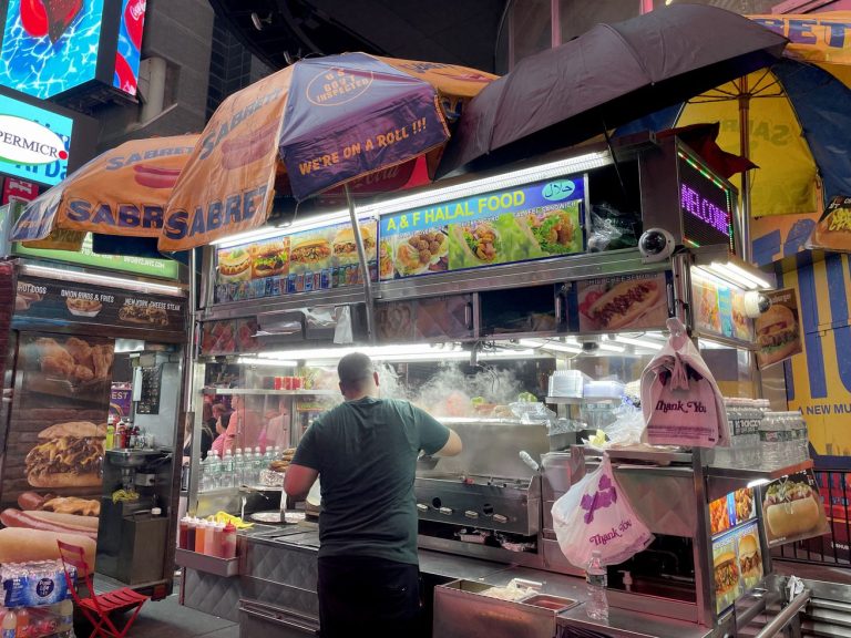 GVL A street food vendor prepares food at a brightly lit halal cart in New York at night, with umbrellas overhead and colorful signs displaying menu items. Smoke rises from the grill as the vendor cooks under neon city lights, highlighting vibrant food carts.