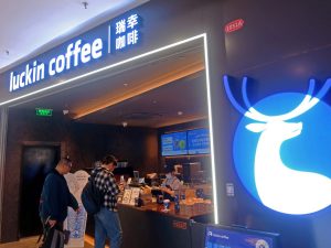 GVL Two people stand at the counter of Luckin Coffee’s first U.S. cafe, featuring a bright blue sign with a white deer logo. Menu boards and staff are visible inside the modern, well-lit café of China’s biggest coffee chain.