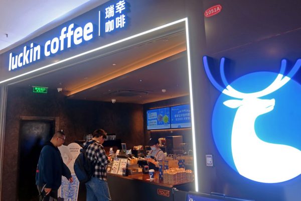 GVL Two people stand at the counter of Luckin Coffee’s first U.S. cafe, featuring a bright blue sign with a white deer logo. Menu boards and staff are visible inside the modern, well-lit café of China’s biggest coffee chain.