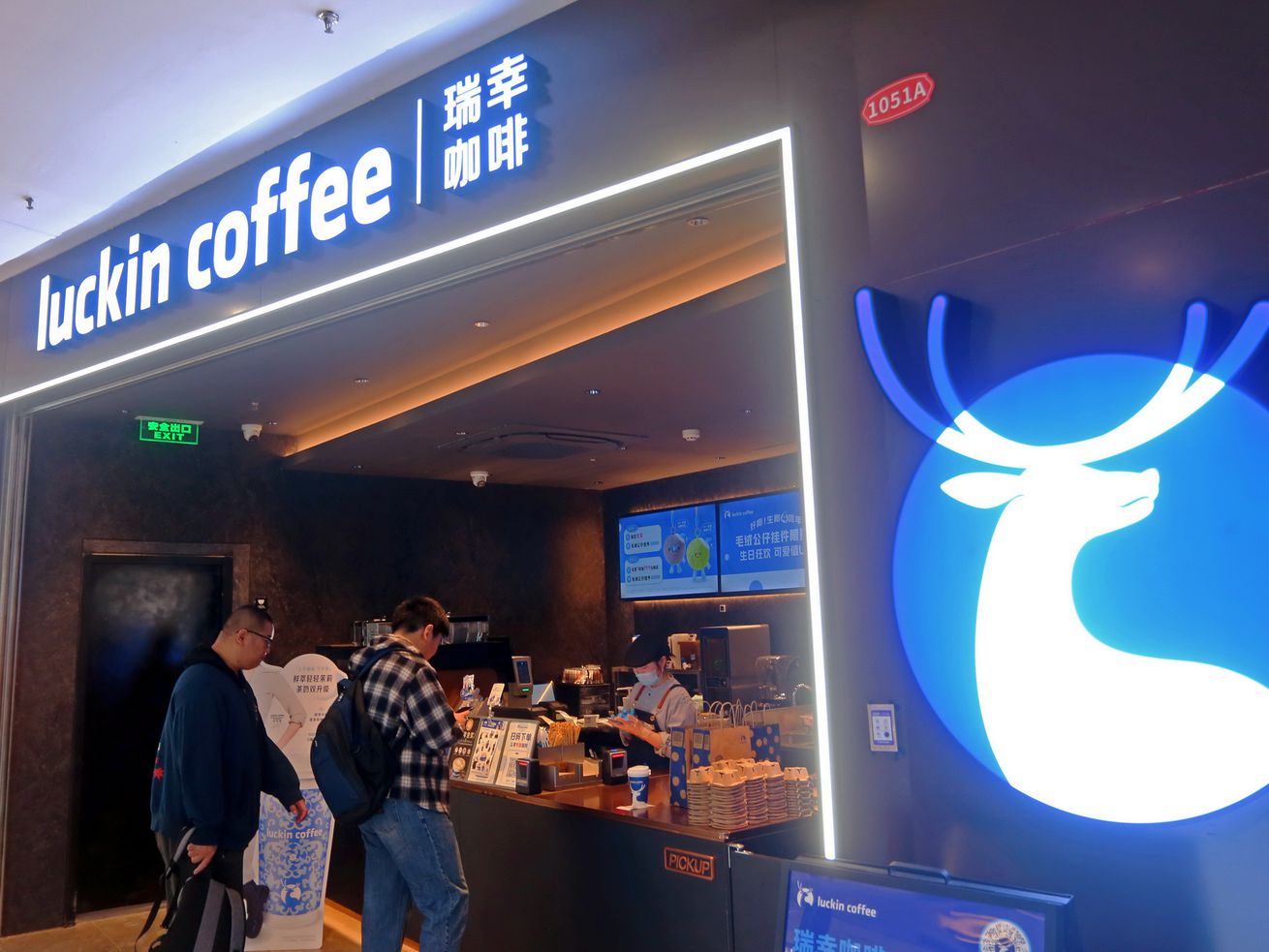 GVL Two people stand at the counter of Luckin Coffee’s first U.S. cafe, featuring a bright blue sign with a white deer logo. Menu boards and staff are visible inside the modern, well-lit café of China’s biggest coffee chain.