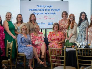 GVL A group of women in colorful dresses pose and smile together indoors at a Make-A-Wish SC fundraising event, with a screen behind them displaying the message, Thank you for transforming lives through the power of a wish.