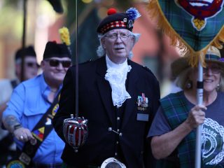 GVL An older man in Scottish attire, including a kilt, jacket, and feathered hat, leads the 2025 Great Scot Parade. He holds a ceremonial sword as others in tartan clothing and sashes walk beside him with banners—perfect for photos and video.