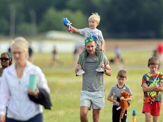 GVL A man carries a young boy on his shoulders while walking outside at the Take Flight 5K, 2025, at Greenville Downtown Airport. The boy holds toys, including Sonic the Hedgehog, as other children and adults enjoy the grassy area with trees in the background.