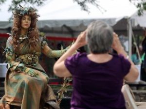 GVL A person dressed as a woodland statue with leaves and vines poses calmly for photos, while another in a purple shirt captures the scene at Artisphere 2025.