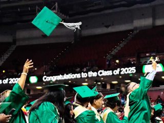GVL Graduates in green caps and gowns toss their caps in the air during the Berea Class of 2025 graduation ceremony. A digital sign in the background reads, Congratulations to the Class of 2025.