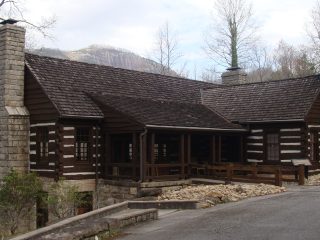 GVL A rustic log cabin with a stone chimney sits on a slight hill at Table Rock State Park, surrounded by bare trees, with a mountain visible in the background under a cloudy sky—a scene worthy of Dennis Chastains Field Notes.
