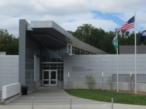 GVL A modern building with gray siding and large windows, flanked by three flagpoles displaying the American flag and two other flags, sits under a cloudy sky—an inspiring site at Greenville Technical College, renowned for its workforce development.