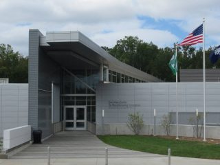 GVL A modern building with gray siding and large windows, flanked by three flagpoles displaying the American flag and two other flags, sits under a cloudy sky—an inspiring site at Greenville Technical College, renowned for its workforce development.