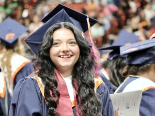 GVL A smiling Carolina Class of 2025 graduate in a navy blue cap and gown stands among fellow graduates at a commencement ceremony, holding papers. The background is filled with more graduates and a blurred audience, capturing the spirit of graduation photos.